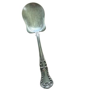 May include: A silver-toned metal serving spoon featuring a wide, rounded bowl and a decorative handle with intricate embossed scrollwork and leaf-like patterns. The metal surface displays a muted, aged finish with subtle variations in color.