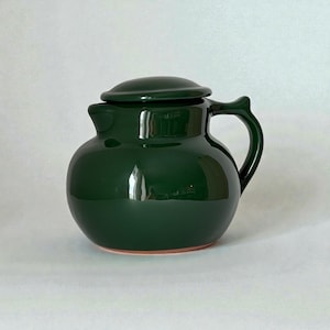Op de afbeelding: Een donker bosgroene keramische theepot met een rond, bolvormig lichaam, een korte tuit en een bijpassend deksel. De theepot heeft een glanzende, reflecterende afwerking, een gebogen handvat aan de rechterkant en een zichtbare terracotta voet.