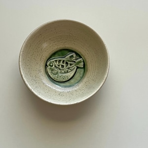May include: A white ceramic bowl with a green, embossed pufferfish design. The bowl has a speckled glaze.