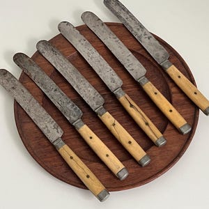 May include: Set of seven antique knives with wooden handles. The knives have a rustic, worn look and are arranged on a wooden platter.