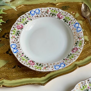 以下が含まれることがあります： 金色の花柄の縁取りが施された白い磁器のスープボウル。縁取りには青いリボン、ピンクのバラ、緑の葉が描かれています。ボウルは金色と緑色のトレイに乗っています。