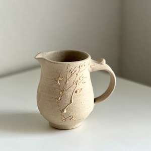 Puede incluir: Una jarra de cerámica beige con asa y un diseño floral en un tono beige más claro.