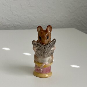 Puede incluir: Una pequeña figura de ratón marrón leyendo un periódico titulado "The Tailor & Cutter". El ratón está posado sobre una bobina de hilo rosa y amarillo, creando una escena en miniatura detallada.