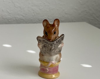 May include: A small, brown mouse figurine reading a newspaper titled "The Tailor & Cutter." The mouse is perched on a pink and yellow spool of thread, creating a whimsical and detailed miniature scene.