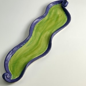 May include: A handcrafted ceramic decorative tray with a wavy, elongated shape. The tray features a vibrant green interior, a deep purple border, and a curled end. The tray is approximately 18 inches long.