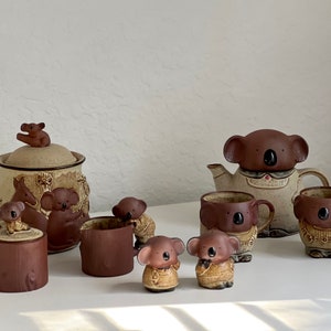 Puede incluir: Un juego de tazas de té y tetera de cerámica marrón y beige con diseños de osos koala. La tetera tiene una tapa de oso koala y las tazas tienen caras de oso koala. También hay un pequeño tarro de cerámica con una tapa de oso koala y una pequeña taza de cerámica.