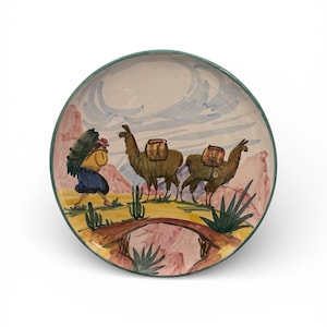 Puede incluir: Plato de cerámica pintado a mano con borde verde. El plato presenta una escena colorida de una persona que lleva un bulto, dos llamas, un puente y elementos del paisaje desértico en tonos rosa, marrón y azul.