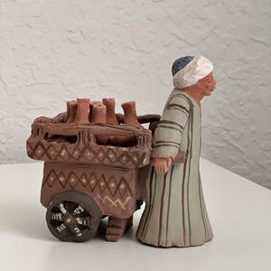 May include: A hand-painted ceramic figurine of a person pushing a cart with clay pots. The cart has a brown and white geometric pattern. The person is wearing a green and white striped robe and a blue and white hat.