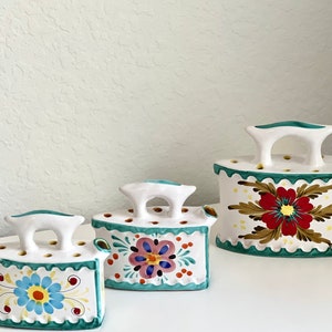 May include: Three decorative ceramic iron-shaped containers. Each is white with colorful floral designs, a turquoise scalloped edge, and a white handle. The largest iron features a red flower, while the others have blue and pink floral patterns.