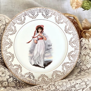 May include: A white porcelain plate with a gold rim and a delicate floral pattern. The center of the plate features a painting of a young girl in a white dress and red hat. The text "Baby Lawrence" is printed below the image.