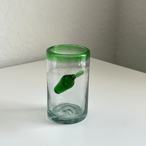 May include: A clear glass shot glass with a green rim and a green glass chili pepper inside.