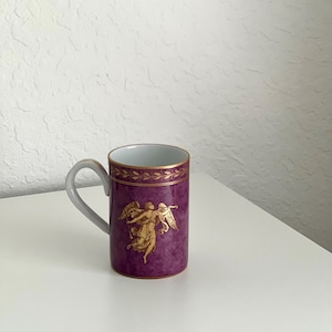 May include: A purple ceramic mug with a gold trim and a gold angel design. The mug has a handle.