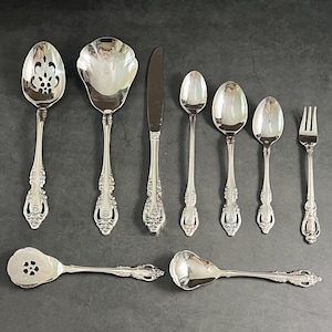 May include: A collection of ornate silver-colored serving utensils, including spoons, a knife, and a fork, arranged on a dark surface. The spoons vary in size and design, with decorative handles and intricate patterns. The knife has a smooth blade.