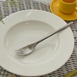 May include: A white ceramic bowl with a silver fork on a white wicker surface. The bowl is empty and the fork is lying on the bottom of the bowl.