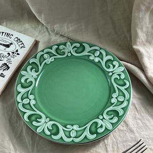 May include: A green ceramic plate featuring a white, decorative, scrollwork design around the rim. A fork and knife with cream handles are in the foreground. A book with the words "Ting Creek" is partially visible.