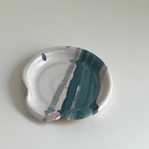 Peut inclure: Un repose-cuillère en céramique blanche avec un motif abstrait bleu et gris. Le repose-cuillère a un bord ondulé et une légère indentation pour tenir les cuillères.