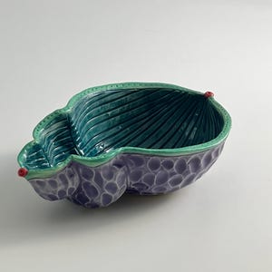 May include: A ceramic decorative bowl in the shape of a leaf. The interior is teal with ribbed detailing, while the exterior is a textured purple. The rim is green, and small red accents are on the edges. The bowl is on a white background.