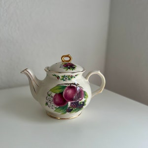 Puede incluir: Una tetera de cerámica blanca con tapa y asa doradas. La tetera está decorada con un patrón floral y un diseño de ciruelas y moras.