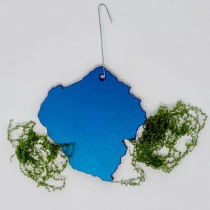 May include: A bright blue, irregularly shaped ornament with a metallic finish and a small hole at the top for hanging. A thin silver metal hook is inserted through the hole. Two clusters of textured, vibrant green moss flank the blue ornament.