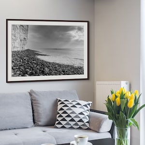 May include: A black and white photograph of a rocky beach with a cliff in the background. The photo is framed in a dark wood frame and is hanging on a white wall above a grey sofa.