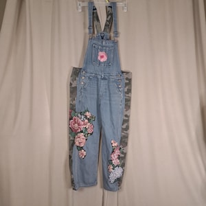 Könnte beinhalten: Hellblaue Jeans-Latzhose mit Gap-Markenetikett, verstellbaren Schulterträgern und einer Brusttasche. Das Kleidungsstück ist mit gestickten Blumenapplikationen in Rosa, Weiß und Grün verziert und hat seitliche Einsätze aus Camouflage-Stoff.