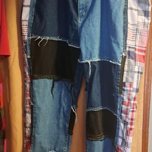 May include: Patchwork denim shorts with a unique design. The shorts feature a mix of blue denim, black denim patches, and red and white plaid fabric. The shorts are hanging on a white hanger.