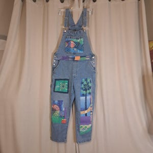 May include: Light blue denim dungarees featuring a bib pocket with a colourful fish graphic, a waist band with orange and purple accents, and various rectangular fabric patches on the legs depicting a sea turtle, palm trees, and abstract patterns.
