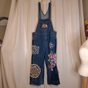 May include: Dark blue denim dungarees featuring a bib pocket with a floral embroidery, a black and white patterned fabric patch, a large beige circular crochet doily appliqué, and a vibrant floral bouquet embroidery on the lower left trouser leg.