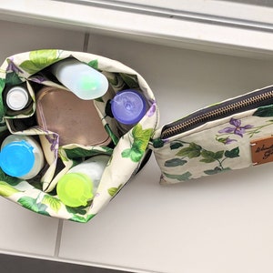 May include: A white and green floral print zipped pouch with a brown leather patch that says "Wanderlust Gear". The pouch is open and filled with various toiletries, including a blue and white bottle, a clear bottle, a blue lidded container, and a green lidded container.