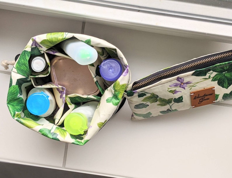 May include: A white and green floral print zipped pouch with a brown leather patch that says "Wanderlust Gear". The pouch is open and filled with various toiletries, including a blue and white bottle, a clear bottle, a blue lidded container, and a green lidded container.