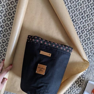 May include: A black fabric bucket bag with a floral print lining. The bag has a brown leather patch with the text "Wanderlust Gear" on it. The bag is wrapped in brown paper.