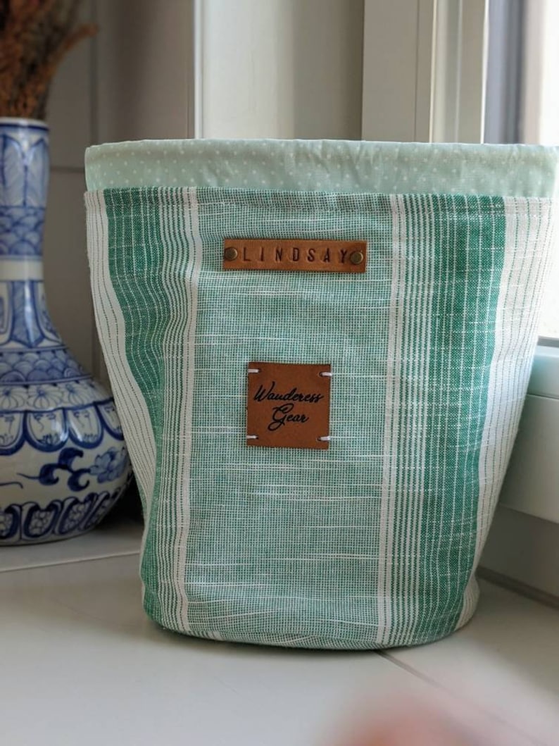 May include: Green and white striped fabric storage bin with a brown leather label that says "Wanderess Gear" and a smaller brown leather label that says "LINDSAY".