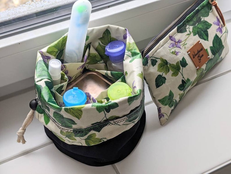 May include: A black and white drawstring bag with a green and white floral pattern. The bag is filled with various small containers and a white tube. The bag is sitting on a white surface. The bag has a label that says "Wanderlust Girl".