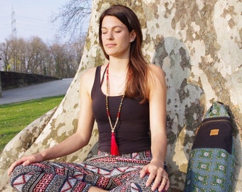 May include: A woman sits cross-legged in front of a tree, wearing a black tank top, patterned pants, and a beaded necklace with a red tassel. She has her eyes closed and appears to be meditating. A yoga mat bag with a patterned design is beside her.