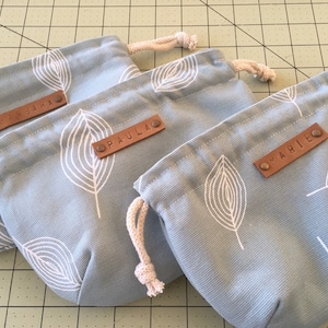 Puede incluir: Tres bolsas con cordón de color azul claro con un estampado de hojas blancas. Cada bolsa tiene una etiqueta de cuero marrón con un nombre: "UNSARA", "PAULA" y "MARIE". Las bolsas tienen un cierre de cordón beige.