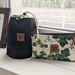 May include: Two black and white fabric pouches with a zipper closure. The pouches have a brown leather patch with the text "Wanderlust Gear" on them. One pouch has a floral print with green leaves and purple flowers. The pouches are on a white surface.