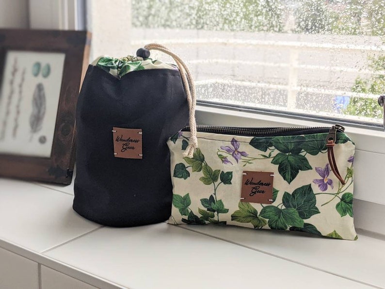 May include: Two black and white fabric pouches with a zipper closure. The pouches have a brown leather patch with the text "Wanderlust Gear" on them. One pouch has a floral print with green leaves and purple flowers. The pouches are on a white surface.