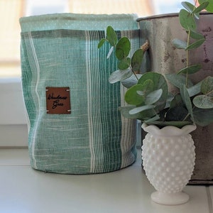 May include: A green and white striped fabric storage bin with a leather label that says "Wanderlust Gear". The bin is sitting on a white surface next to a white ceramic vase with a textured surface. A sprig of eucalyptus is in the vase.