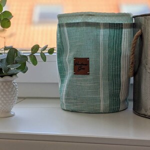 May include: A green and white striped fabric storage bin with a brown leather tag that says "Wanderlust Goods". The bin has a rope handle and is sitting on a white surface next to a metal container.