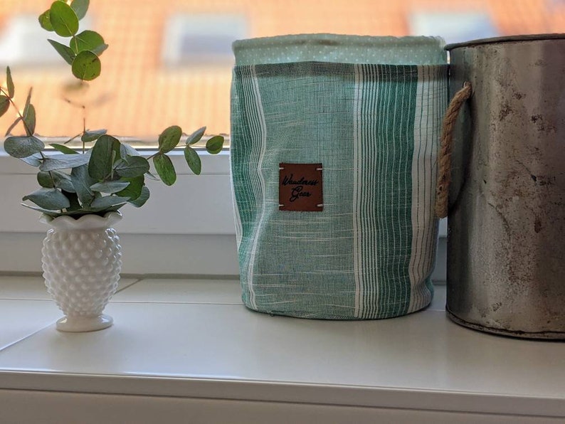 May include: A green and white striped fabric storage bin with a brown leather tag that says "Wanderlust Goods". The bin has a rope handle and is sitting on a white surface next to a metal container.