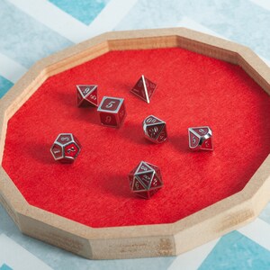 May include: A wooden octagonal tray with a red felt interior holds several red and silver dice. The dice are of various shapes, including a d4, d6, d8, d10, d12, and d20. The tray sits on a blue and white patterned surface.