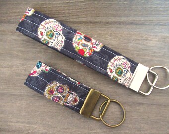 May include: Two black and white keychains with a colorful sugar skull design. The larger keychain has a silver key ring and the smaller keychain has a gold key ring.