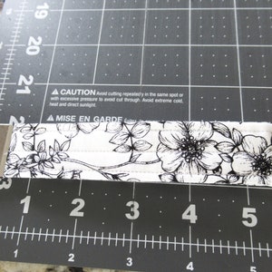Op de afbeelding: Een witte stoffen sleutelhanger met een zwart bloemenpatroon en een zilveren sleutelring. De sleutelhanger is ongeveer 7,6 cm lang. De tekst op de sleutelhanger luidt: "▲ CAUTION Avoid cutting repeatedly in the same spot or with excessive pressure to avoid cut through. Avoid extreme cold, heat and direct sunlight. ▲ MISE EN GARDE &Eacute;vitez"