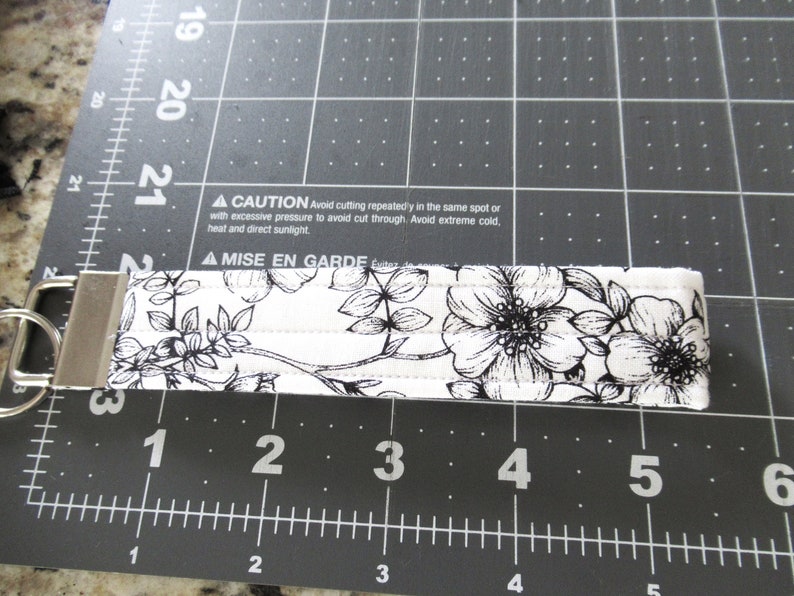 Op de afbeelding: Een witte stoffen sleutelhanger met een zwart bloemenpatroon en een zilveren sleutelring. De sleutelhanger is ongeveer 7,6 cm lang. De tekst op de sleutelhanger luidt: "▲ CAUTION Avoid cutting repeatedly in the same spot or with excessive pressure to avoid cut through. Avoid extreme cold, heat and direct sunlight. ▲ MISE EN GARDE &Eacute;vitez"
