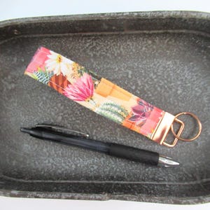 May include: A pink and orange fabric key chain with a floral print of cacti and flowers. The key chain has a gold metal key ring.