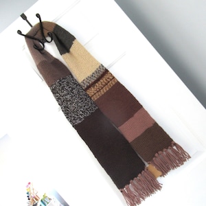 May include: A multi-colored knitted scarf with a variety of brown, tan, and gray sections. The scarf has a fringed edge and is hanging on a white door. The scarf is made of a soft, warm material.