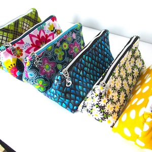 May include: A collection of six colorful zippered pouches. The pouches feature various patterns, including floral, plaid, and polka dots. Each pouch has a silver zipper and a contrasting lining. The pouches are arranged in a row, showcasing their different designs.