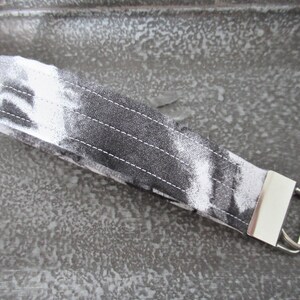 Op de afbeelding: Een sleutelhanger van wit en zwart stof met een zilveren metalen ring. De stof heeft een tie-dye patroon.
