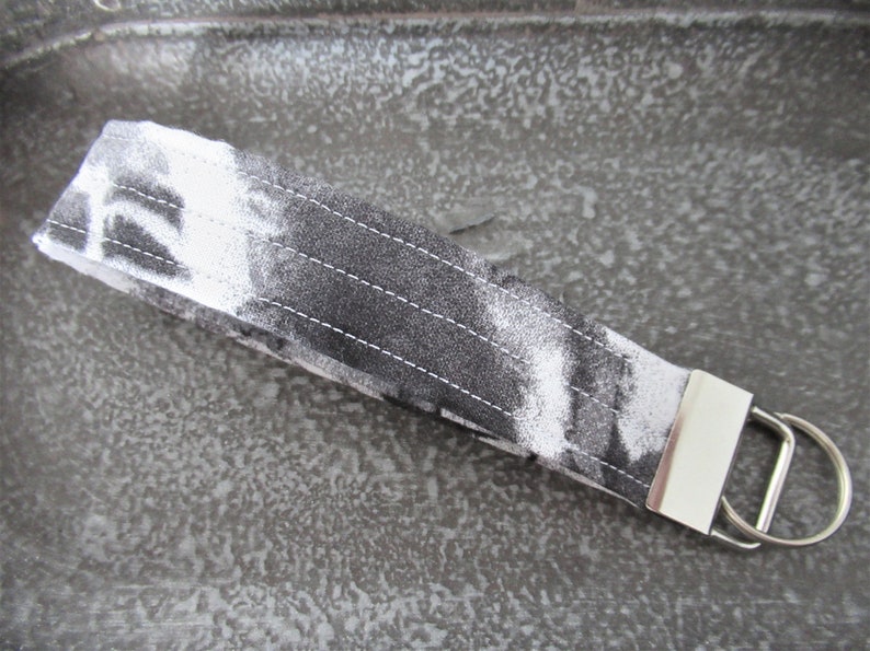 Op de afbeelding: Een sleutelhanger van wit en zwart stof met een zilveren metalen ring. De stof heeft een tie-dye patroon.