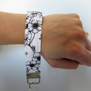 Op de afbeelding: Een witte sleutelhanger van stof met een zwarte bloemenprint en een zilveren sleutelring.